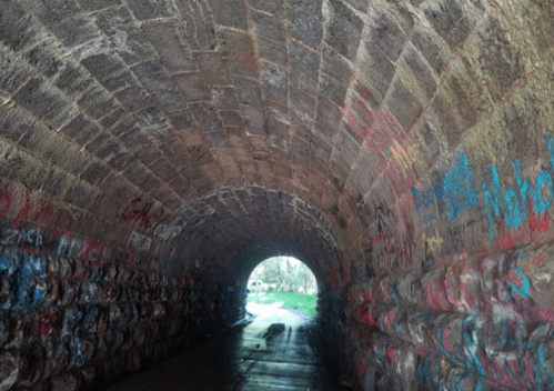 A stone tunnel with colorful graffiti on the walls, leading to a bright exit at the far end.
