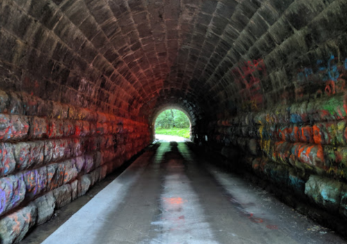 A stone tunnel with colorful graffiti, leading to a bright opening at the end, surrounded by greenery.