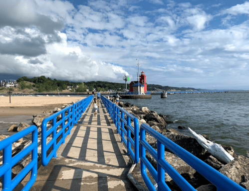 A blue pier extends over water, leading to a red lighthouse, with people walking and a sandy beach in the background.