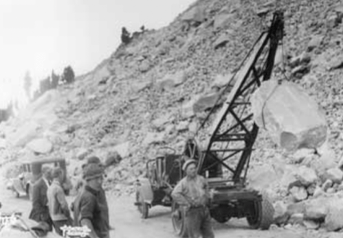 A historic scene showing a crane lifting a large rock, with workers and vehicles nearby on a rocky hillside.