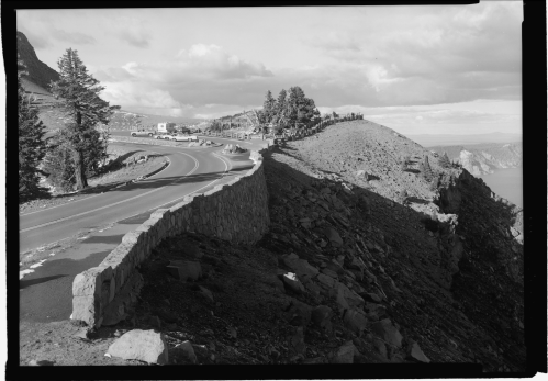 A winding road curves along a rocky cliff with trees and a distant view of a lake under a cloudy sky.
