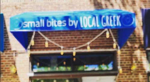 A blue awning with the text "small bites by LOCAL GREEK" above a restaurant entrance, adorned with string lights.