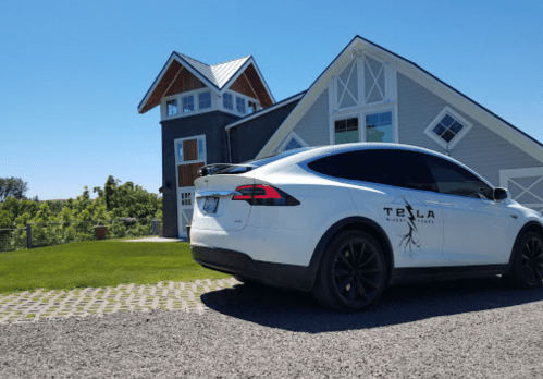 A white Tesla Model X parked in front of a modern house on a sunny day, with green grass and a clear blue sky.