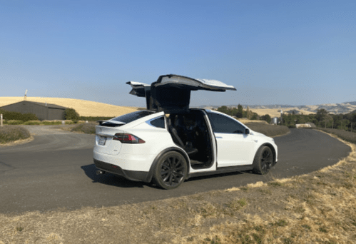 A white Tesla Model X parked on a winding road, with its falcon-wing doors open, surrounded by dry grass and hills.