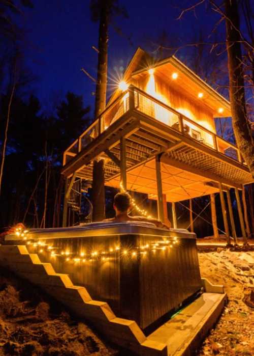 A cozy treehouse illuminated at night, with a hot tub adorned with string lights in the foreground.