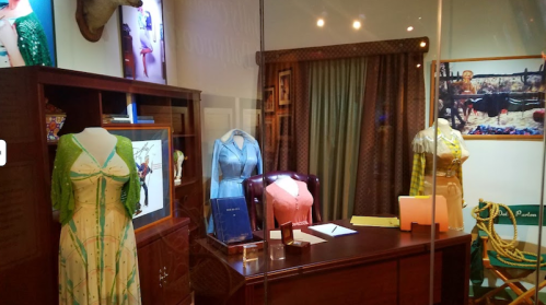 A display of vintage dresses and a desk in a glass case, showcasing fashion and decor in a stylish room.