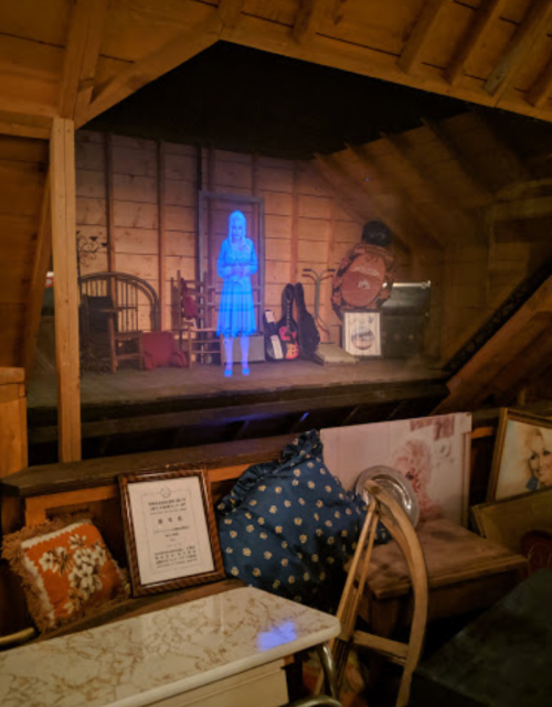 A dimly lit attic with a holographic figure of a girl in a blue dress, surrounded by vintage furniture and decor.