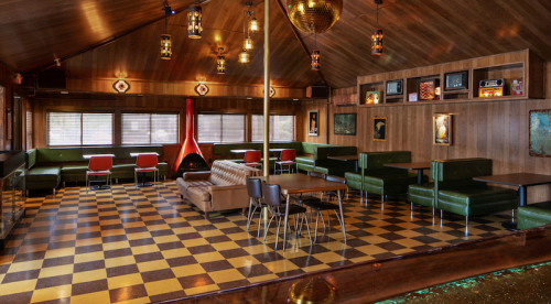 A cozy, retro-style lounge with wooden walls, checkered floor, green seating, and a fireplace.
