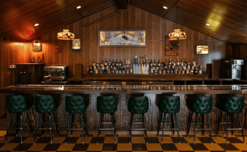 A rustic bar with a wooden counter, green upholstered stools, and shelves lined with various bottles. Warm lighting enhances the ambiance.