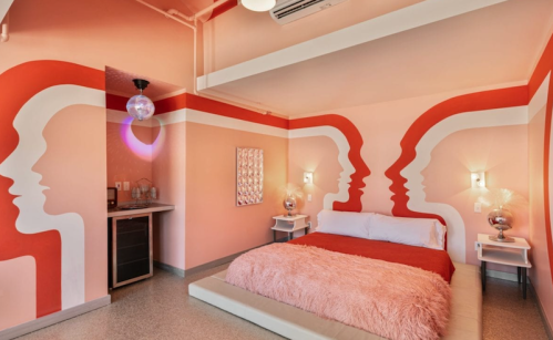 A stylish bedroom with pink walls featuring abstract face designs, a plush bed, and modern decor elements.