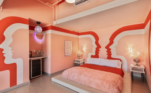 A stylish bedroom with pink walls featuring abstract face designs, a plush bed, and modern decor elements.