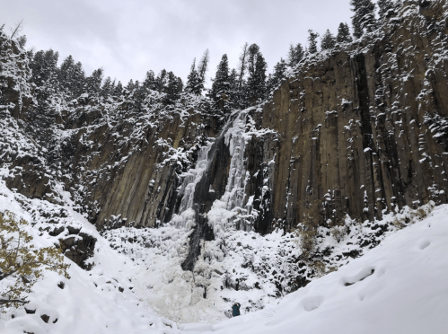 A snowy landscape featuring a frozen waterfall cascading down rocky cliffs, surrounded by evergreen trees.
