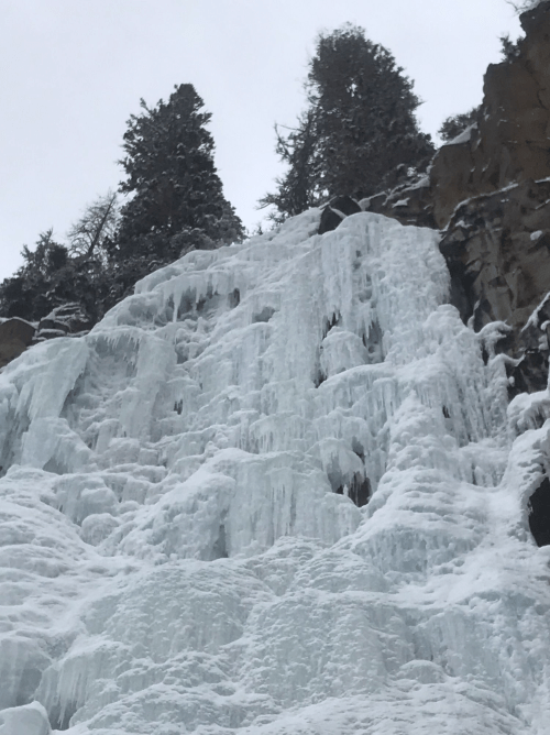 A frozen waterfall cascading down a rocky cliff, surrounded by snow-covered trees.
