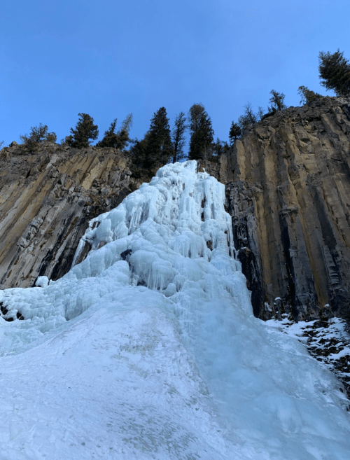 A frozen waterfall cascading down rocky cliffs, surrounded by evergreen trees under a clear blue sky.