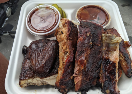 A takeout container with grilled ribs, two sauce cups, and pickles on the side.