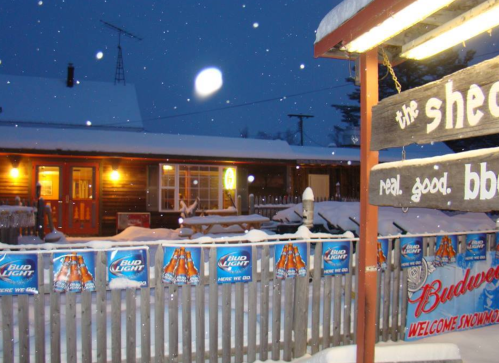 A cozy cabin in the snow at night, with festive beer banners and a welcoming sign. Snowflakes are falling gently.