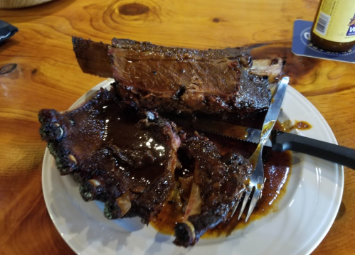 A plate of succulent, glazed beef ribs with a knife and fork resting beside them on a wooden table.