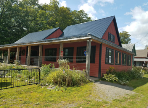 A red house with a metal roof, surrounded by greenery and a gravel path, under a partly cloudy sky.