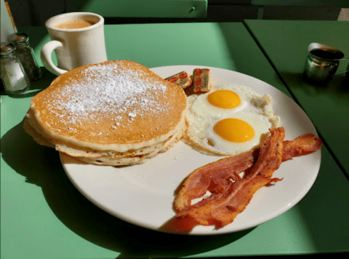 A plate of pancakes with powdered sugar, two sunny-side-up eggs, crispy bacon, and a cup of coffee on a green table.