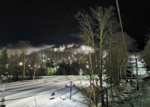 A snowy landscape at night, featuring ski slopes, trees, and distant lights from a ski resort.