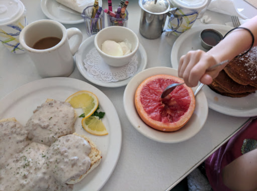 A breakfast table with pancakes, eggs Benedict, a grapefruit, coffee, and cream. A hand is scooping grapefruit.