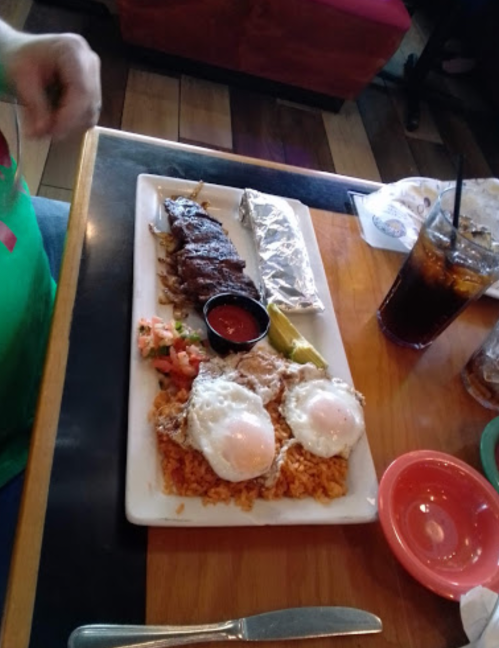 A plate of rice topped with two fried eggs, grilled meat, a foil-wrapped item, and a side of salsa and vegetables.