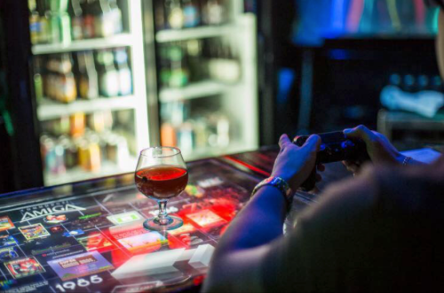 A person holds a game controller while sitting at a table with a drink, surrounded by colorful bar lighting.