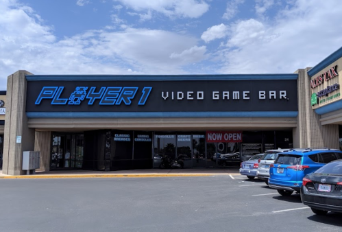 Exterior of "Player 1 Video Game Bar" with a blue sign, cars parked outside, and a clear sky above.