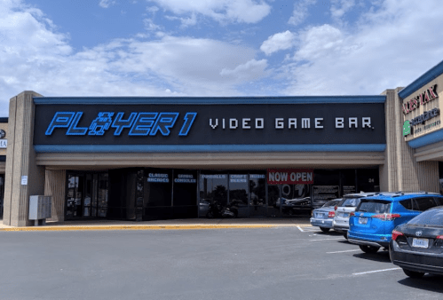 Exterior of "Player 1 Video Game Bar" with a blue sign, cars parked outside, and a clear sky above.