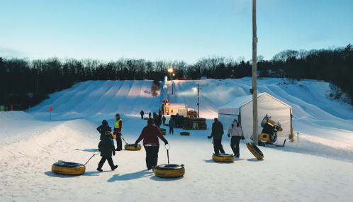 People carrying snow tubes at a winter tubing park, with illuminated slopes and a tent in the background.