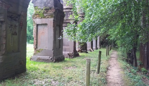 Ruins of stone structures covered in moss, surrounded by greenery and a dirt path.