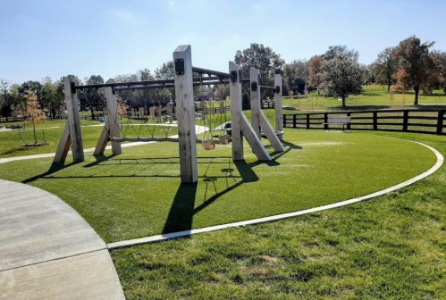 A playground with swings and climbing structures on green grass, surrounded by trees and a clear blue sky.