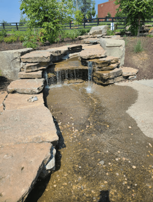 A small stone waterfall flows over rocks into a shallow stream, surrounded by greenery and a pathway.
