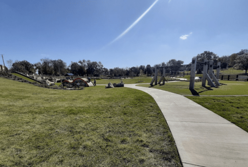 A sunny park scene featuring a winding path, playground equipment, and grassy areas under a clear blue sky.