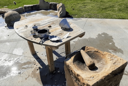 A wooden table with bowls and utensils, next to a stone grinding tool, set in a grassy outdoor area.