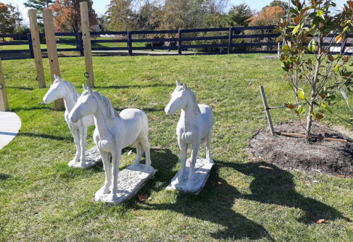 Three white horse statues stand on a grassy area, with trees and a fence in the background.