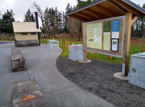A park area featuring a map board, benches, and trash bins, surrounded by trees and a cloudy sky.
