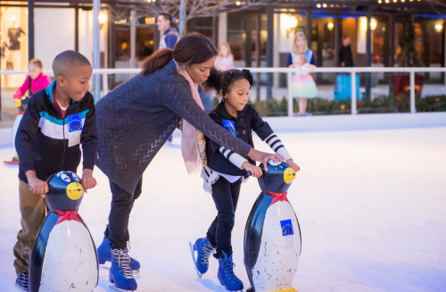 A woman assists two children on ice skates using penguin-shaped skating aids at an outdoor rink.