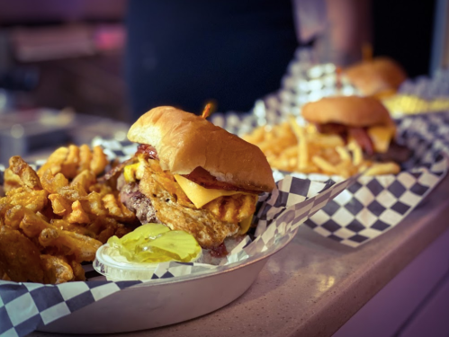 Three delicious burgers with cheese and toppings, served with curly fries and pickles on a checkered tray.