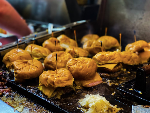 A sizzling grill with multiple cheeseburgers, each topped with a toothpick, surrounded by melted cheese and onions.