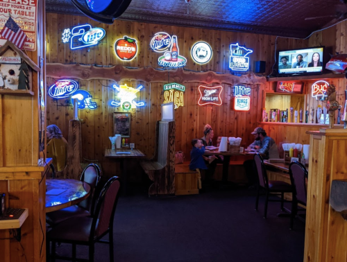 Cozy restaurant interior with wooden walls, neon signs, and two tables occupied by diners. A TV is visible in the background.