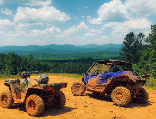 Two muddy ATVs parked on a dirt trail, overlooking a lush green landscape and mountains under a blue sky with clouds.
