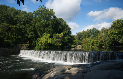 A serene waterfall cascades over rocks, surrounded by lush green trees and a clear blue sky.