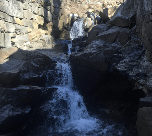 A serene waterfall cascading over rocky terrain, surrounded by stone walls and natural scenery.