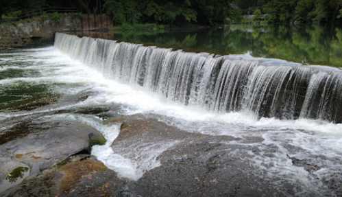 A serene waterfall cascading over a stone ledge into a calm river, surrounded by lush greenery.