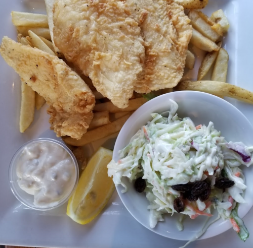 A plate with fried fish fillets, French fries, coleslaw, a lemon wedge, and a small cup of tartar sauce.
