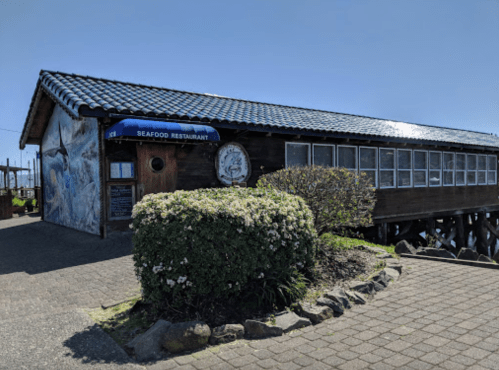 A seafood restaurant with a wooden exterior and blue roof, surrounded by greenery and a paved walkway.
