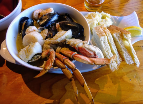 A bowl of mixed seafood including crab, shrimp, and mussels, served with bread and a lemon wedge on a wooden table.
