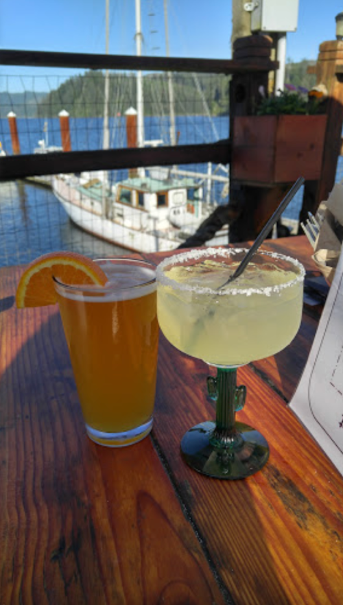 A pint of beer and a margarita with a lime wedge sit on a wooden table by a waterfront. A boat is visible in the background.