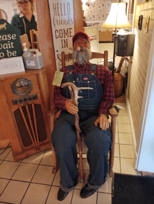 A lifelike mannequin of an elderly man in overalls and a plaid shirt, sitting in a rocking chair with a cane.